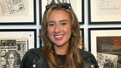 Stacey Burns, who has long brown hair with blonde highlights stands in front of a series of picture frames on a wall. She has sunglasses on her head and is wearing a black leather jacket.