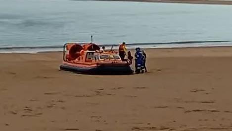 An orange RNLI hovercraft beached on a north Norfolk sandy beach. The grainy footage shows lifeboat crew helping someone on to the beach while a cluster of HM Coastguard staff in blue and white stand next to the boat.