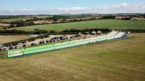 The giant green and white water slide in a large field. There is cars parked in the car park next to it.