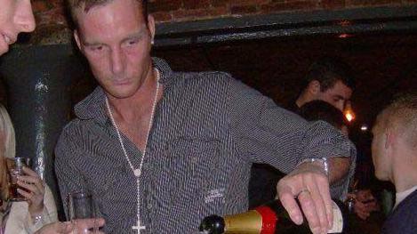 Adam Minett pours a bottle of champagne with his left hand. He is wearing an open-necked shirt with a chain around his neck, and is in the middle of a crowd in a bar in Liverpool. 