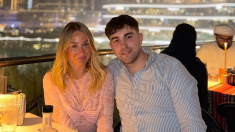 Caitlyn, a 21-year-old woman from Blackpool, is sitting in a bar on a roof terrace with the Dubai skyline behind them