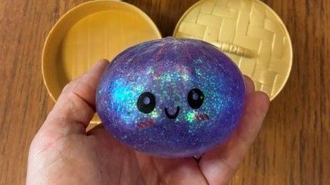 One of the squeezy dumpling toys being held in a hand. It is dumpling-shaped and made of sparkly purple plastic, with black eyes and a small mouth. It comes with gold-coloured plastic steamer