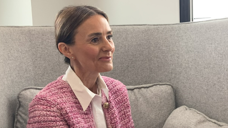 Leyla Yildirim is sitting on a grey sofa. She is wearing a pink blazer and a pink shirt. She has her brown hair neatly tied back and she is smiling.