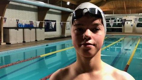 Theo standing in front of the pool wearing a white swimming cap and black goggles on his head