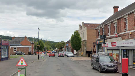 A long straight road with shops and buildings on either side. There are cars parked on the right hand side and cars driving down the road.