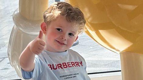 Peter Maughan is standing outside on a summer's day wearing a grey T-shirt with the Burberry logo in red capital letters. He is making a thumbs-up sign with his head tilted and he is smiling at the camera looking happy. He has short, blond hair and is face is flushed.