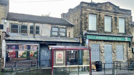 A boarded-up heritage building, bearing the name Netherton Village Hall, stands behind a bus stop with abandoned shops to its left.