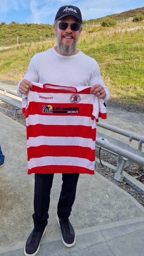 Matt Damon is wearing a baseball cap, a pair of shades and a white long-sleeved top. He has blue jeans and trainers. He is standing outside and is holding a red and white striped Bonnyrigg Rose top.