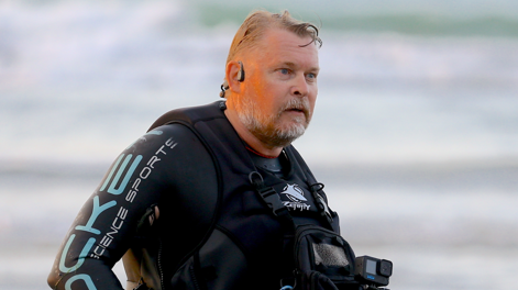 A man wearing a wetsuit and an earpiece stands in front of the sea on a beach.