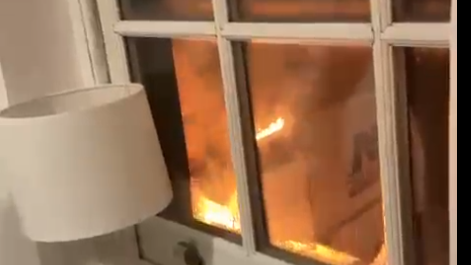 Grainy video still of a large van on fire with yellow and orange flames coming through its doors. It is pictured through a bedroom window, with a lamp visible on the windowsill.