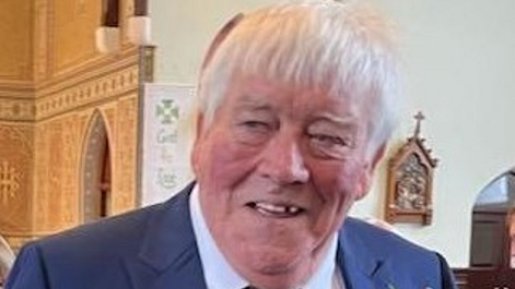 An elderly man with short white hair, a blue jacket and white shirt, smiles while he stands in a church.