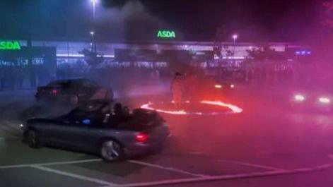 Cars drive around a "ring of fire" in an Asda supermarket car park in Bletchley, Milton Keynes. The picture is blurry, although you can see people standing in the middle of the ring and others watching on.