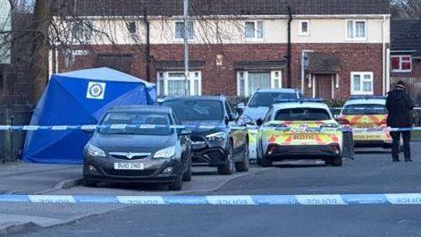 A residential street that is cordoned off with police tape. There are two police cars in the background and an officer standing, with their back to the camera, near the tape