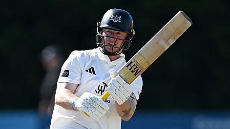 Miles Hammond pulls the ball away through mid-wicket. He is holding his bat high to the right of the picture. He is wearing his whites and has a dak blue helmet on
