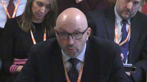 A bald man in black glasses and a black suit with white shirt and black tie. He is sat looking forwards to a person off to the left of the camera. Two people in suits are sat behind him, also facing forwards