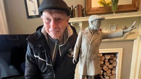 A man wearing a flat cap and black Port Vale branded coat poses next to a miniature version of a statue of himself.