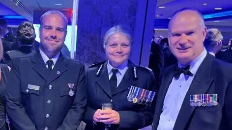 Justin Robbie, Sarah Crew, and Mark Shelford, at an awards ceremony, smiling. The three are wearing a varying number of medals, and Sarah is clutching a flute of champagne.