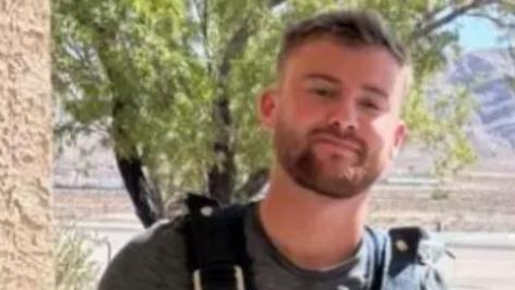 Mitchell Deakin who has light brown hair and a brown beard and wears a grey t-shirt. He is wearing parachute equipment and in the backdrop is a small aircraft on a landing strip.