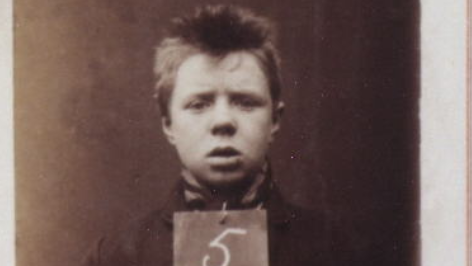 A mugshot of a young male prisoner., John Porter He is baby-faced with a mop of brown hair and is holding a sign with a 5 on it - the rest of the sign is cut off. The image is in sepia tones, and was taken on an old film camera. 