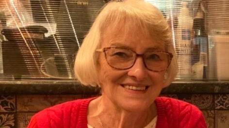 Andrea Sloan is seated indoors at what appears to be a restaurant or café. She is wearing a bright red, cable-knit cardigan over a white textured top, accessorised with a delicate necklace featuring a small pendant. The background includes patterned ceramic tiles along the wall.