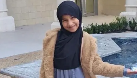 Inaayah Makda, wearing a brown furry jacket and a headscarf, smiles at the camera as she stands next to a water fountain in the shape of a star.