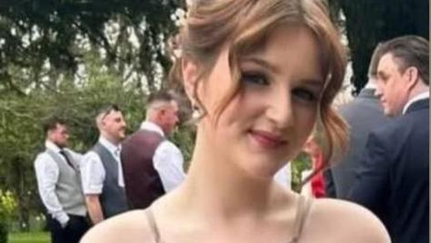 Lainie is wearing a beige dress and holding a bunch of dried flowers. She smiling at the camera. Her brown hair is tied up with the two front pieces down and curled. It seems she is a bridesmaid at a wedding.