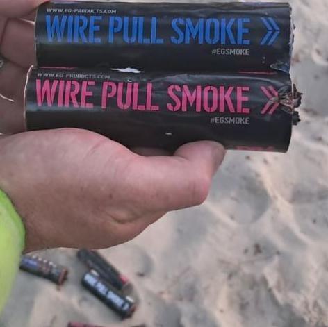 Hands, with the cuff of a high-vis jacket visible, holding tubes of smoke bombs, with a number of discarded tubes on the sand below.