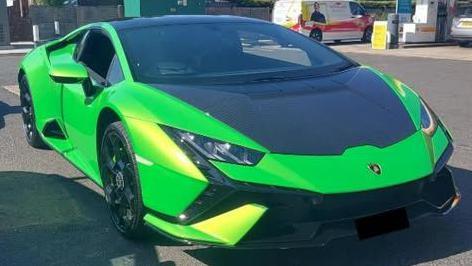 A green Lamborghini, parked up in a petrol station. It is green with black bits on its hood. A van is in the distance.
