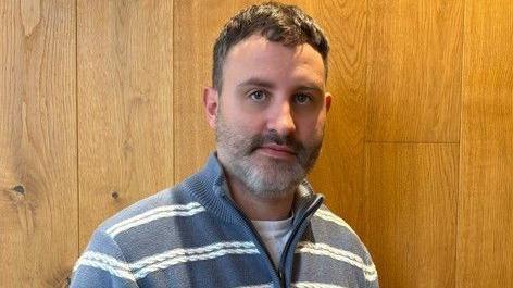 Dominic Webb, who has short brown hair and a beard, looking at the camera wearing a blue zip-up fleece with white hoops. He is stood in front of wooden panels.