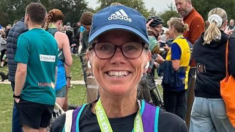 Clare standing outside wearing running gear, with a medal around her neck. It appears she has just completed a running race. She is wearing a blue cap and a black t-shirt. She is smiling at the camera.