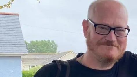 Ian Airlie in a blurry snapshot taken in a doorway. He has a ginger beard, bald head and black-frames glasses. He is wearing a black T-shirt and hads the strap of a rucksack over one shoulder.