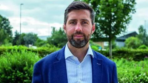 Former South Gloucestershire leader Toby Savage smiling at the camera and wearing a blue jacket and shirt. He is stood in front of green bushes and trees.