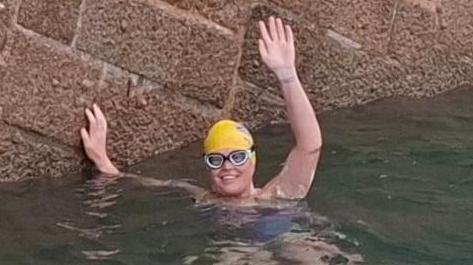 Marie-Helene Sanderson in the water. She is wearing goggles and a yellow swim hat and is waving at the camera while smiling broadly. Her other hand is on a stone sea wall.