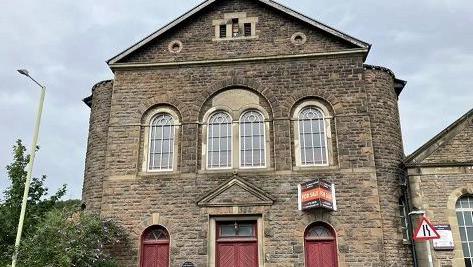 Front facade of the chapel with three double wooden doors side by side painted dark red and four arched windows above. A for sale sign has been attached to the wall at the front.