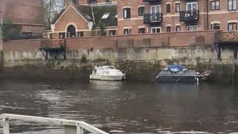A calm riverside scene featuring small docked boats and a large brick residential building with balconies and varied windows. 