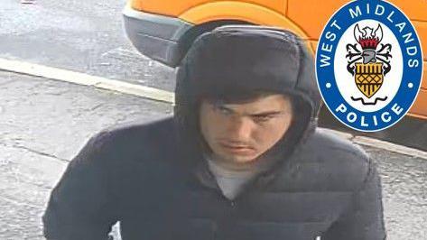 CCTV of a man wearing a dark blue coat with the hood up. A pavement and road can be seen behind him with an orange car parked next to it.