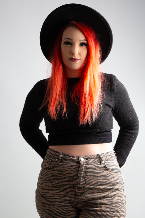 Woman with long orange hair wearing a black hat. She's wearing a black cropped long sleeve top and zebra print brown and black jeans.

