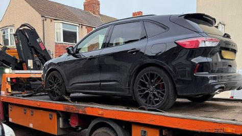 A close-up of a black Ford Kuga with black wheel trims which is been towed away on an orange flat-bed truck. Two-storey terraced houses can just be seen behind the vehicles.