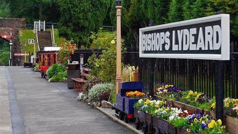 The platform at Bishop's Lydeard with a sign on the right of the shot with the name of the station. There are flowers running along the edge of the platform as well as a shelter further down. In the background are trees surrounding the entire platform.