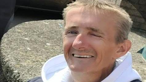 A man wearing a white hoodie smiles for the camera. He has short blond hair. 