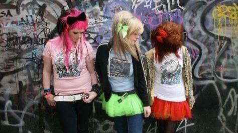 Three girls looking down to the right in front of a graffiti wall. Kerry on the left wears a pink top, bracelets, a white belt and black trousers. She has bright pink and black hair with a fringe. A black bow rests on the top of her head. Kelly in the middle has a blue top with an old boom box pictured on it. She also wears a black sweatshirt, neon green tutu and blue leggings. She has a blonde hair and a fringe, with a matching neon green bow. Debbie on the right is looking down. She has red hair, a red bow and bright red tutu with black tights. She has a white top and leopard print hoodie. 