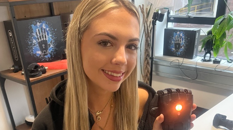 Sadie, who has long blonde hair and is wearing grey tracksuit bottoms, a black top and a black jumper, is holding up the prosthetic hand, which has an orange light flashing from the centre of it.