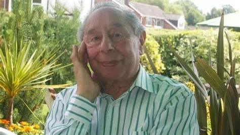 A man sits in a garden. He has grey hair and he is wearing a green striped shirt. Trees can be seen in the background.