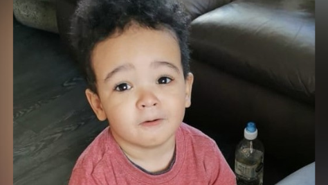 A photo of Daniel Aruebose. He is a young child, around two and a half years old, who is standing near a brown leather sofa. He is wearing a long-sleeved red top and is looking at the camera.