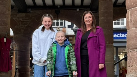 Two children and a woman stand together in a medieval open fronted building. On the left is a 12 year old girl with brown hair in bunches and a pale grey hoodie. Next to her is a seven year old boy on crutches, with no hair, wearing a blue hoodie and a green coat. On his right is a woman with long brown hair and a long purple coat.