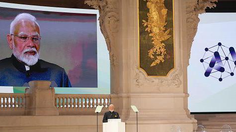 The image shows Narendra Modi, India's prime minister, on the podium speaking at the AI Action Summit in Paris in February 2025. There's also a big screen live streaming his speech right behind the stage where he is standing. 