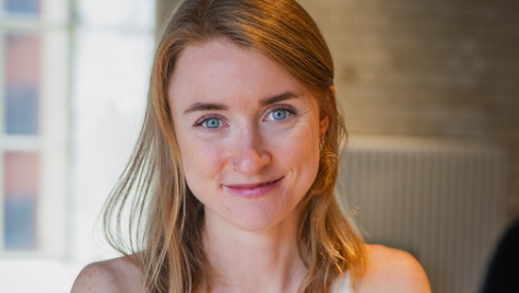 Woman with mouse coloured hair and blue eyes smiling at the camera. She is standing inside a room with a window in the background.