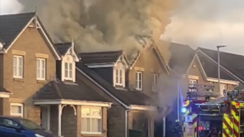 Smoke billowing from a house roof, with a fire engine outside on the street.