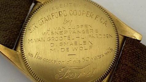 The gold back case of a Rolex watch, which has woven brown fabric straps. The back is inscribed with the words "Presented to Sir Stanford Cooper F.C.A. by J. Van Luppen, W. Snepwangers, M. Van Grootel - L Pardoen, D. Smaelen, M. De Wolf, on the occasion of his 30 years with Ford.