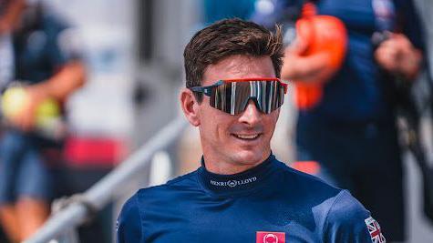 Dylan Fletcher with short dark hair, wearing dark goggles and a blue T-shirt with a Union Jack logo.  He is outside, alongside a railing with people behind him.